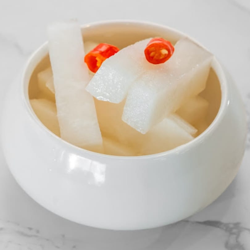 White pickled radish cubes with red chili slices in a white bowl on a marble surface.