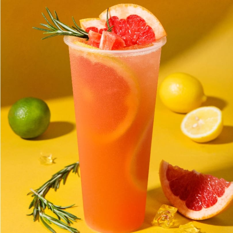 A tall glass of pink grapefruit drink with rosemary, surrounded by citrus fruits on a yellow surface.