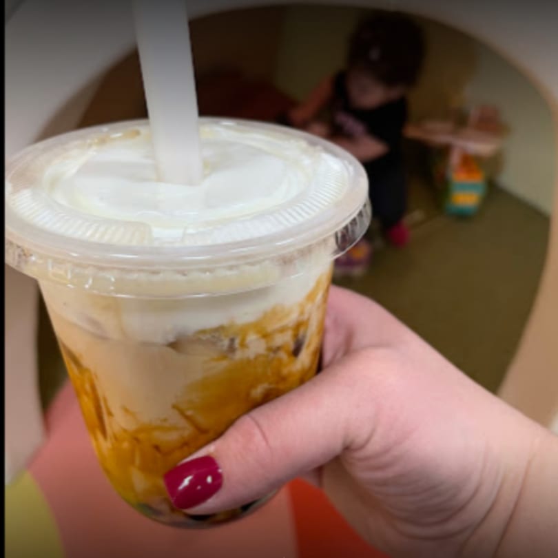Hand holding a Chick n'Cha bubble tea with a child playing in the background.