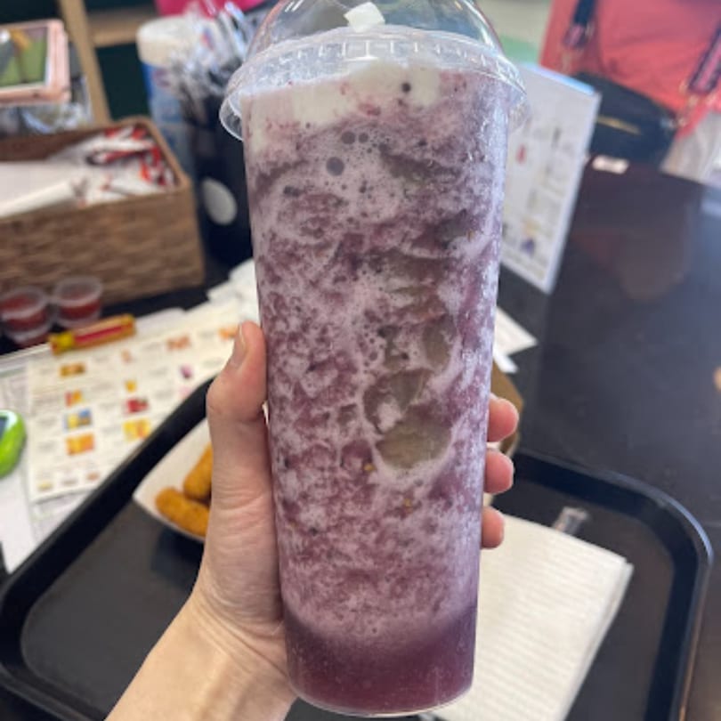 Hand holding a tall plastic cup of purple Chick n'Cha bubble tea with foam, on a tray.