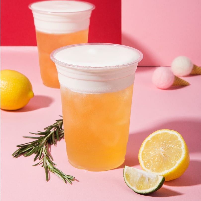 Two Chick n'Cha lemonades with cream tops, lemons, lime, and rosemary on a pink and red background.