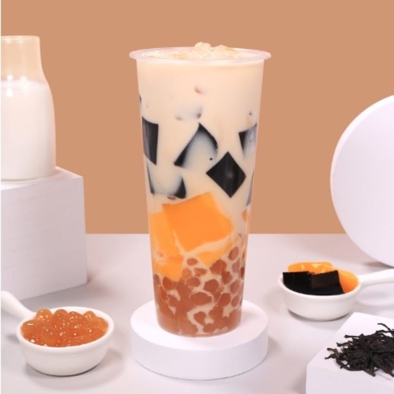 Chick n'Cha bubble tea with black and orange jellies, boba, and milk, surrounded by ingredients.
